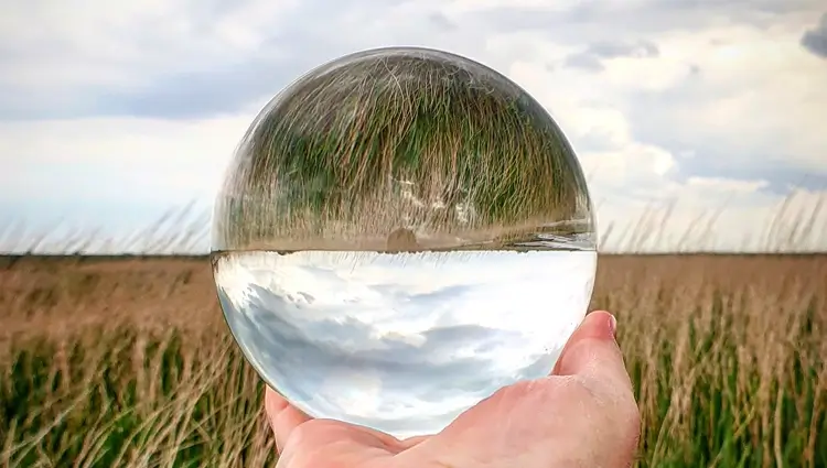 Person hält Glaskugel vor Feld, worin sich Himmel und Feld umgekehrt spiegelt